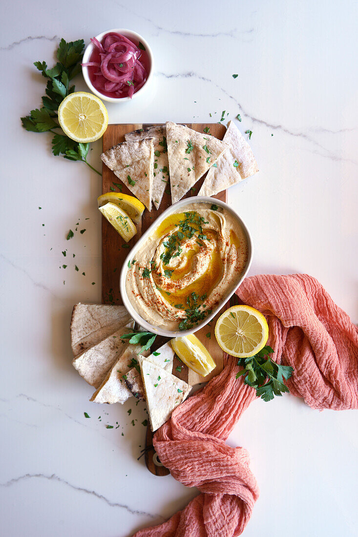 Chickpea hummus served with olive oil, pickled red onion, lemons, and flatbread on a white marble table background. Top view flatlay.