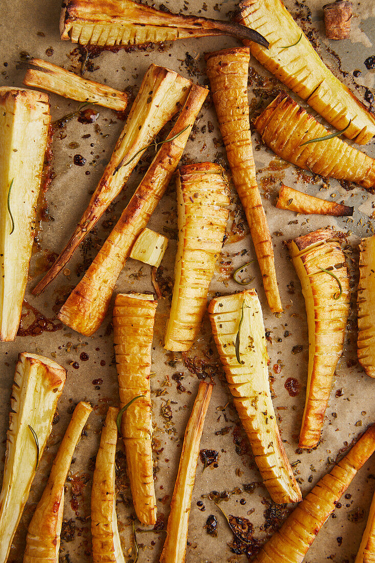 Top view of appetising baked parsnip sticks with rosemary and olive oil on parchment paper in the kitchen while cooking