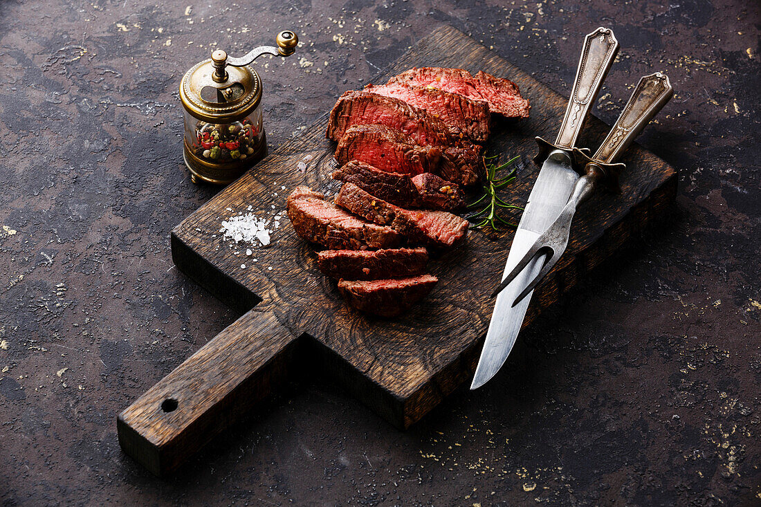 Sliced grilled roast beef steak with knife and fork on a wooden cutting board