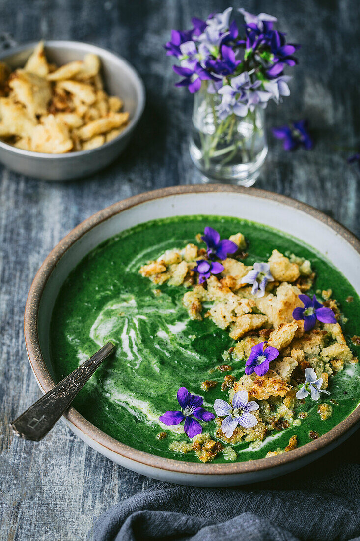 Green vegetable soup with croutons and edible violets in a bowl with spoon and garnishes
