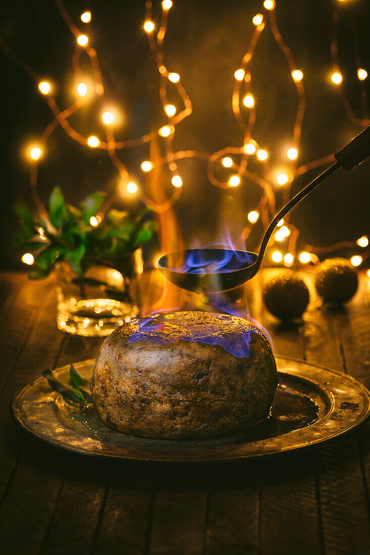 Christmas pudding with spoon and flame on pewter plate with holly and Christmas lights