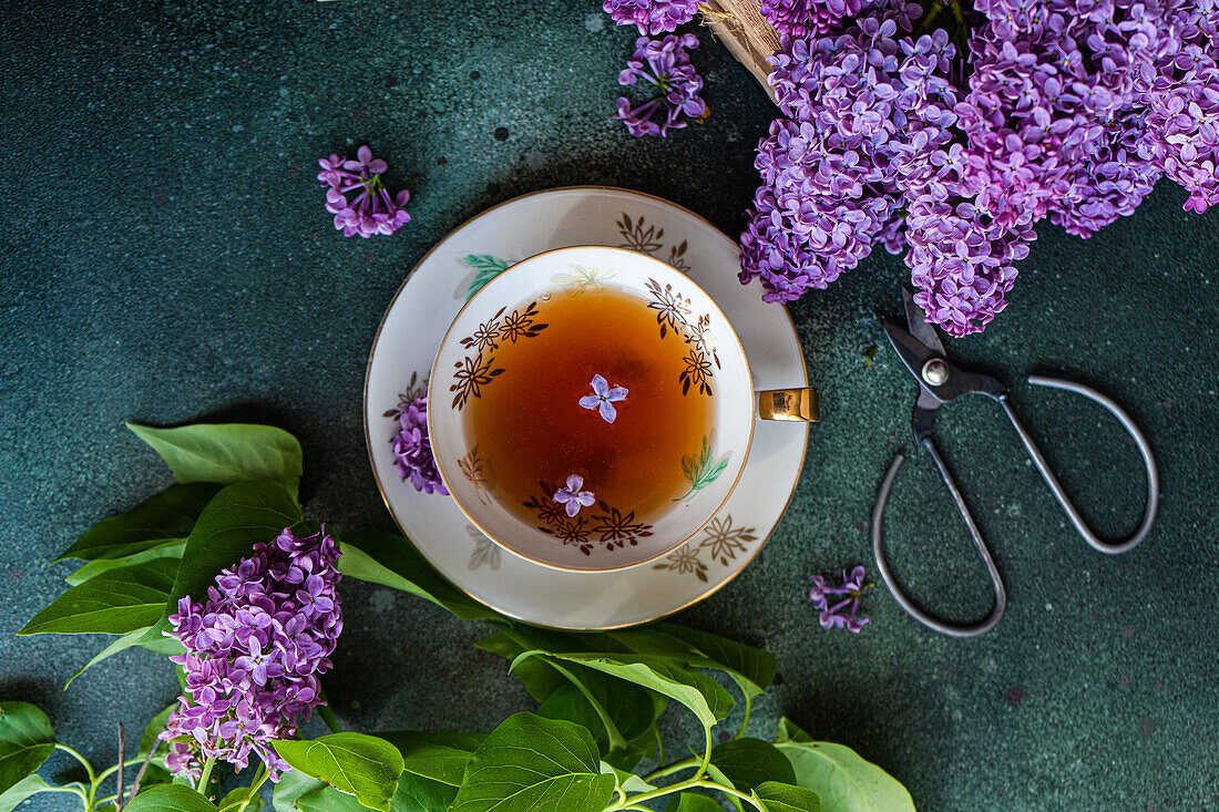 Top view of tasty black tea in vintage white cup on mint green concrete table with aromatic lilac flowers