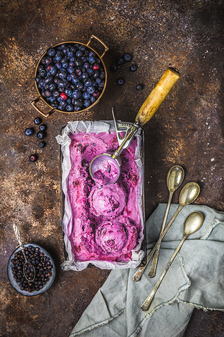 Blueberry ice cream in an old tin with fresh blueberries in a copper strainer, juniper berries in a small bowl and an antique ladle, dessert spoon and linen napkin