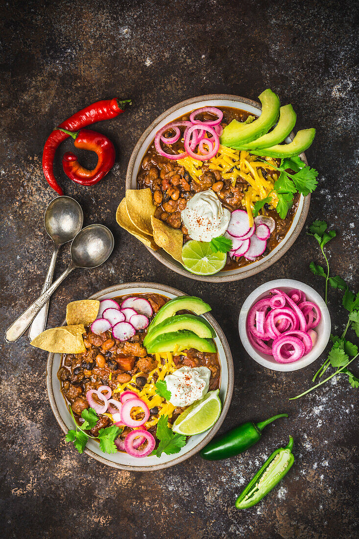 Chili con carne in zwei Schalen mit Beilagen, eingelegten roten Zwiebeln und Löffeln in Blasen auf dunklem Hintergrund