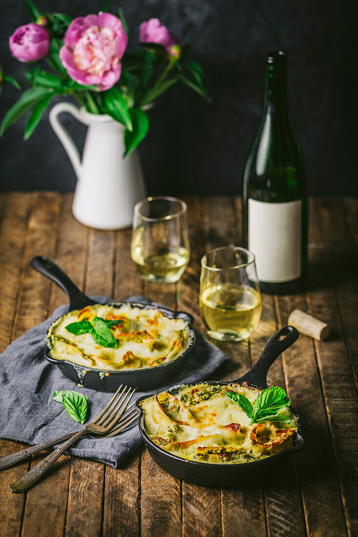 Table setting with baked pasta in two small cast-iron pans with wine glasses, wine bottle and flowers