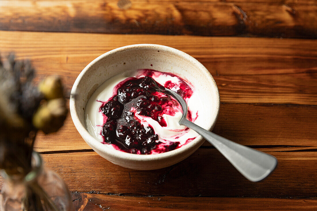 Bowl of yoghurt and blackberry compot on wooden table with silver spoon.