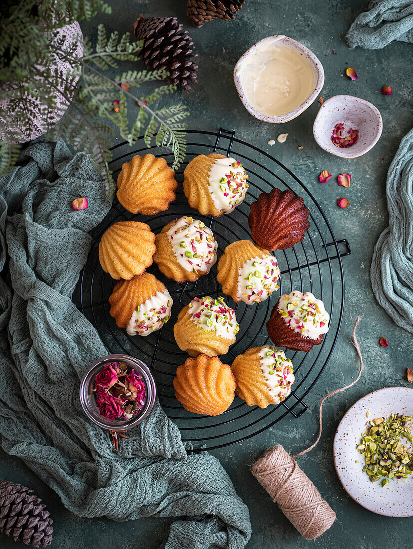 Festliche Madeleines auf einem Kühlgestell, dekoriert mit weißer Schokolade, Pistazien und roten Blütenblättern auf grünem Hintergrund und Tuch