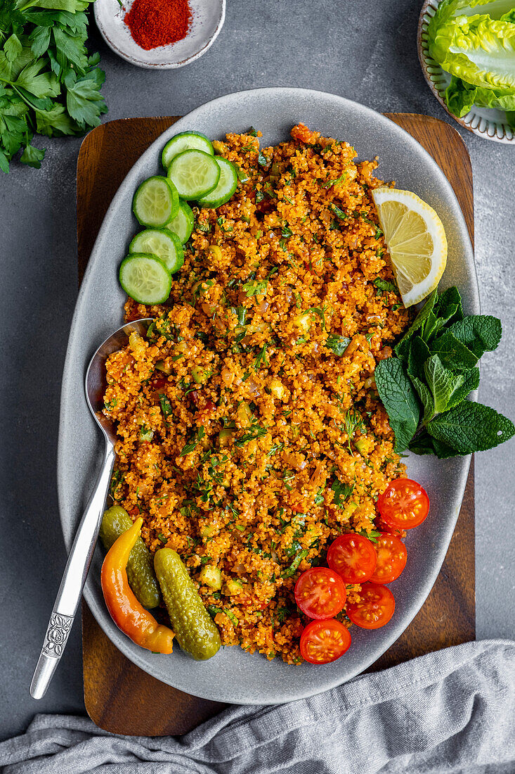 Turkish bulgur salad garnished with tomato and cucumber slices, fresh mint leaves, gherkins and lemon on an oval plate