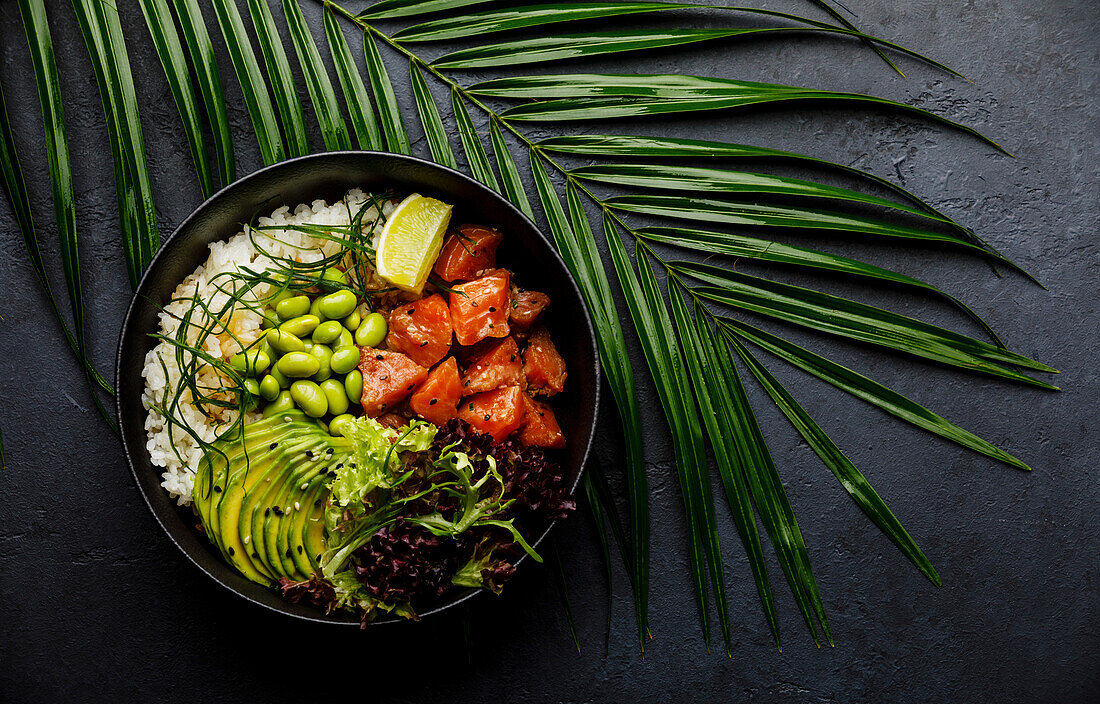 Lachs Poke Bowl Roher Fischsalat Asiatisches Trendgericht mit Sojabohnen, Edamame, Reis, Avocado und Salat in Schale auf tropischem Blatt und dunklem Hintergrund