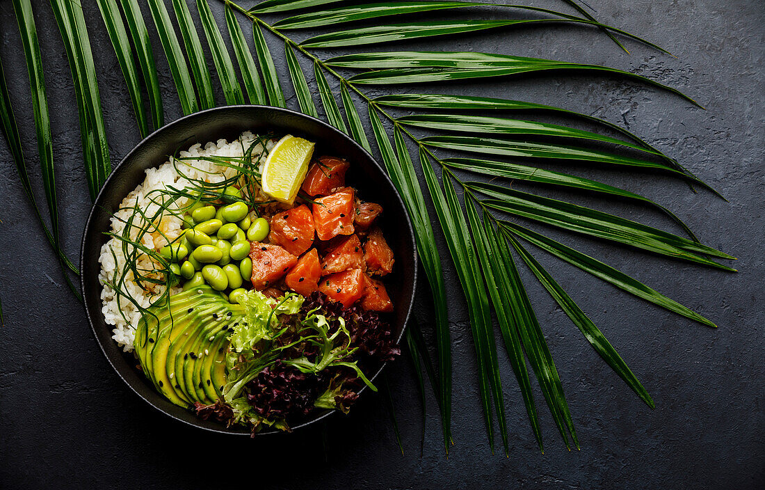 Salmon Poke bowl Raw fish salad Asian trendy food with soy beans edamame, rice, avocado and lettuce in bowl on tropical leaf and dark background