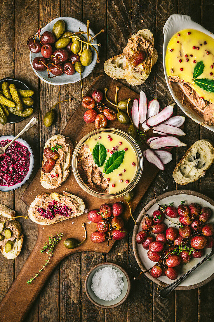 Pâté spread in a glass on a wooden board with roasted grapes, radishes, gherkins and baguette