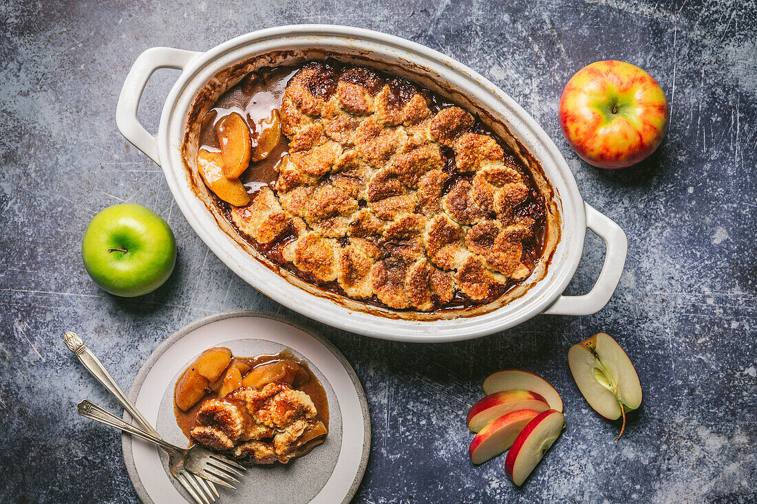Apple Cobbler in ceramic baking dish with serving on plate and fresh apples on blue background
