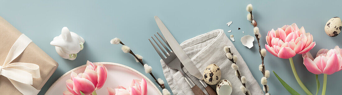 Banner. Gedeckter Tisch. Ein modischer, minimalistischer Teller mit einer Leinenserviette, Messer und Gabel, Geschenkbox, Ostereier, Federn und Tulpen auf blauem Hintergrund. Ansicht von oben