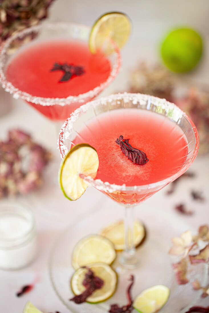 Two hibiscus margartia cocktails garnished with a salm rim and a wheel of dried limes