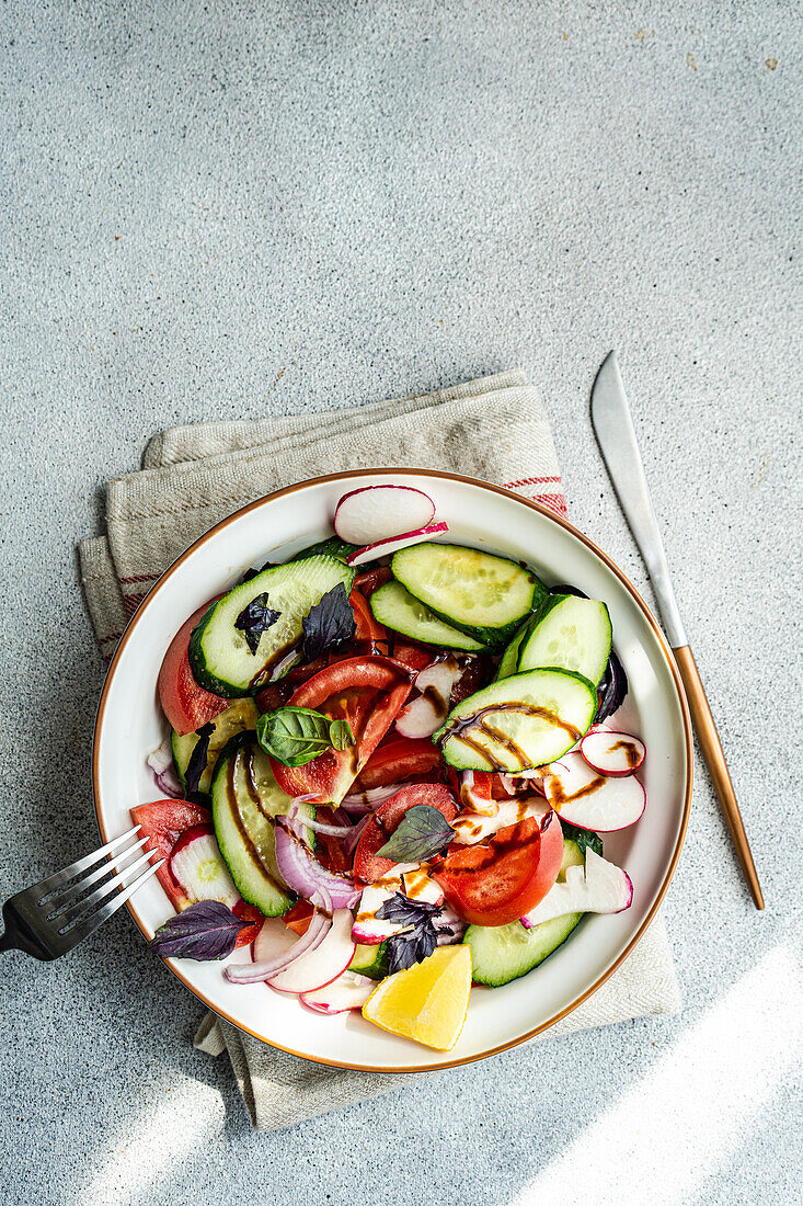 High angle of a delicious vegetable salad with cucumber, lemon, onion and tomato with green leaves and dressing in a bowl placed on the cloth on the table