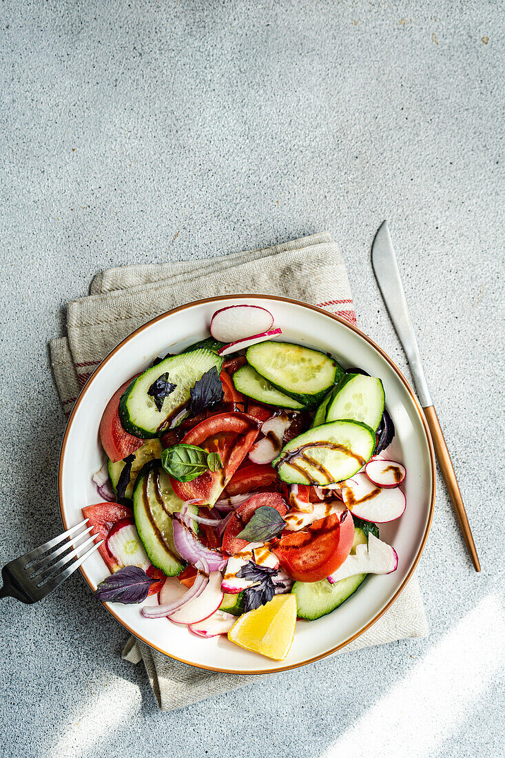 Hoher Winkel eines leckeren Gemüsesalats mit Gurke, Zitrone, Zwiebel und Tomate mit grünen Blättern und Dressing in einer Schüssel, die auf dem Tuch auf dem Tisch steht