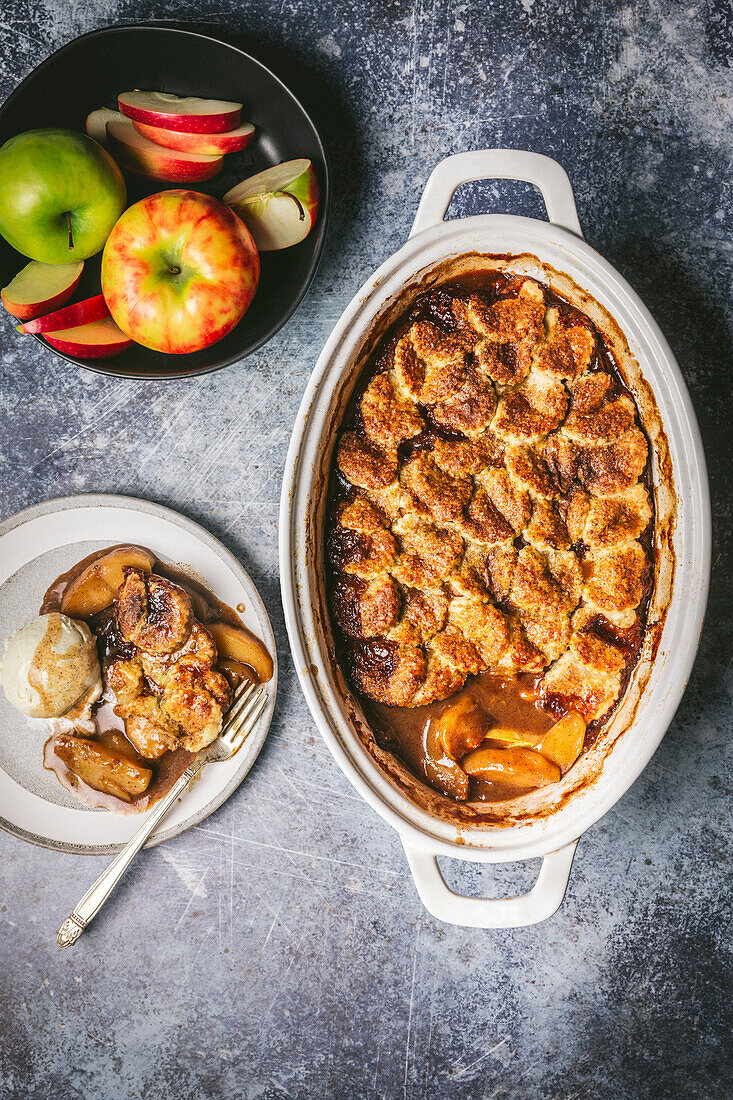 Apple Cobbler in einer Keramik-Backform, serviert auf einem Teller mit Vanilleeis und einer Schale mit frischen Äpfeln