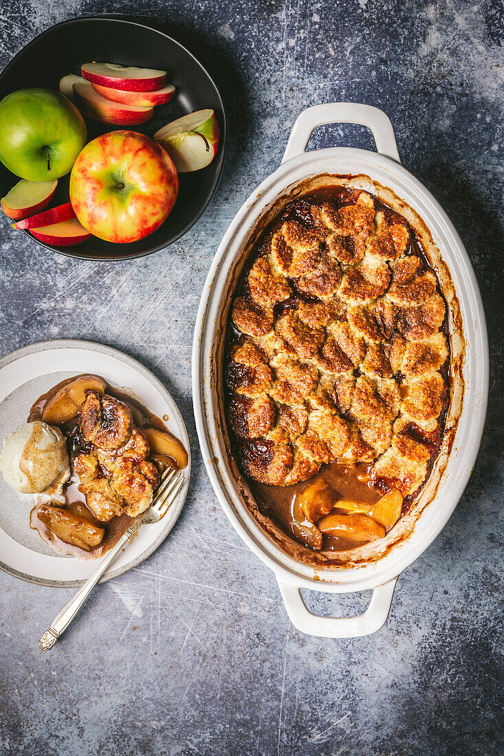 Apple cobbler in a ceramic baking dish, served on a plate with vanilla ice cream and a bowl of fresh apples