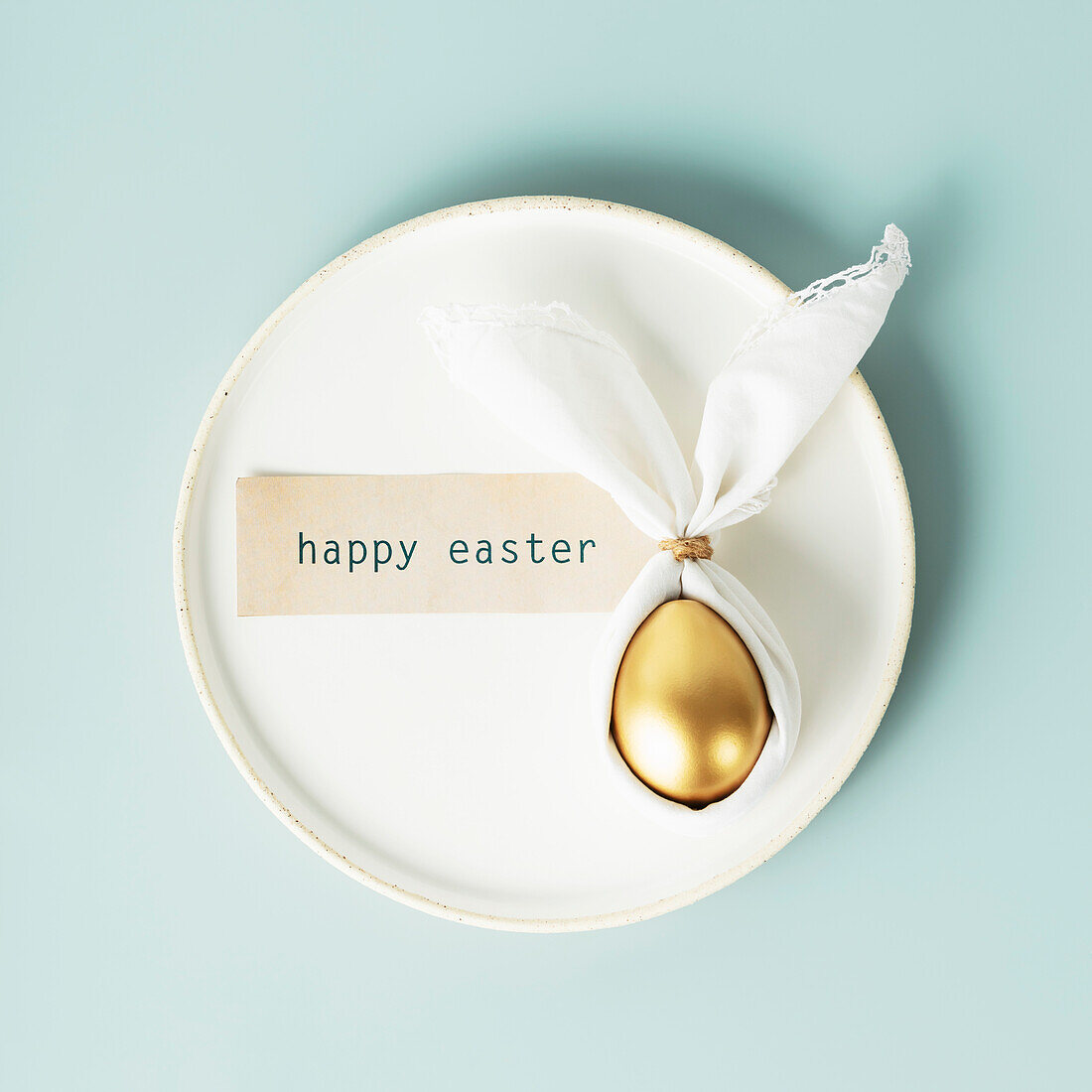 Stylish Easter flat lay with golden egg in Easter bunny napkin on white plate on blue background. Minimalist modern Easter table decorations. Flat lay view from above