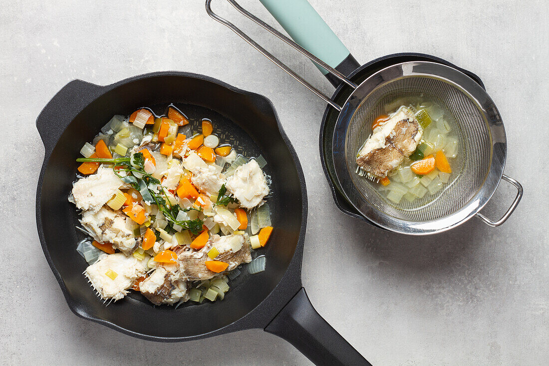Draufsicht auf Bratpfannen mit Gemüse und Fisch in Erdnussöl und Kräutern in Fumet mit Karotten und Trockenfisch auf grauer Fläche in der Küche
