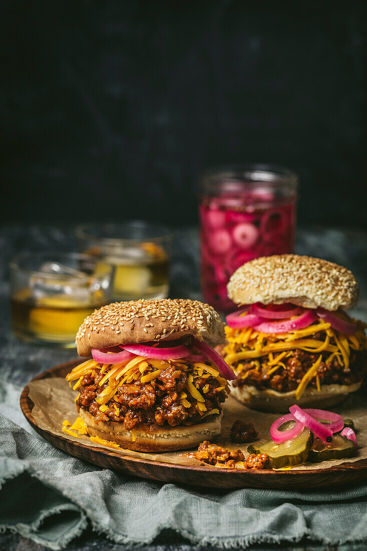 Frontal view of sloppy joes (minced beef sandwiches) with cheddar cheese, pickled red onions on a wooden plate with pickle jar in the background