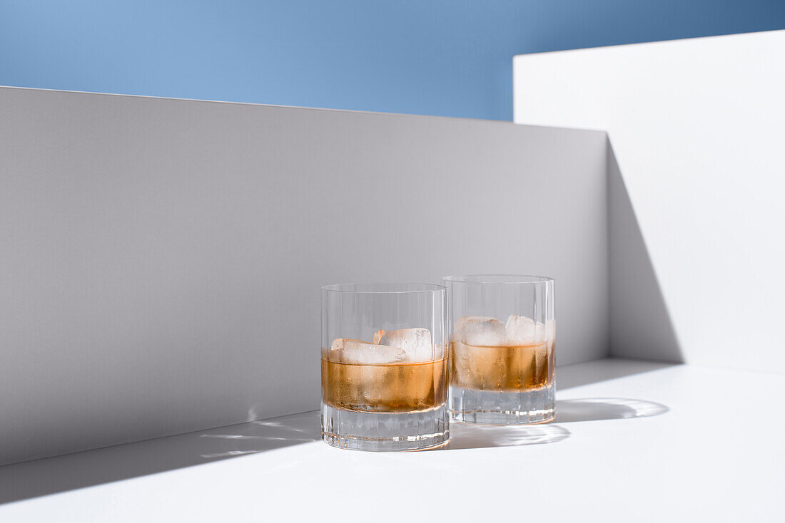 Front view of clear glasses filled with cold, refreshing scotch whiskey with ice cubes on a white surface against a white and blue background