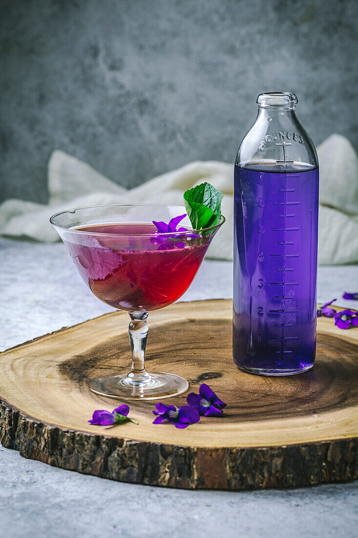 Lila Cocktail im Vintage-Glas mit Blumengarnitur und Sirup auf rohem Holzbrett