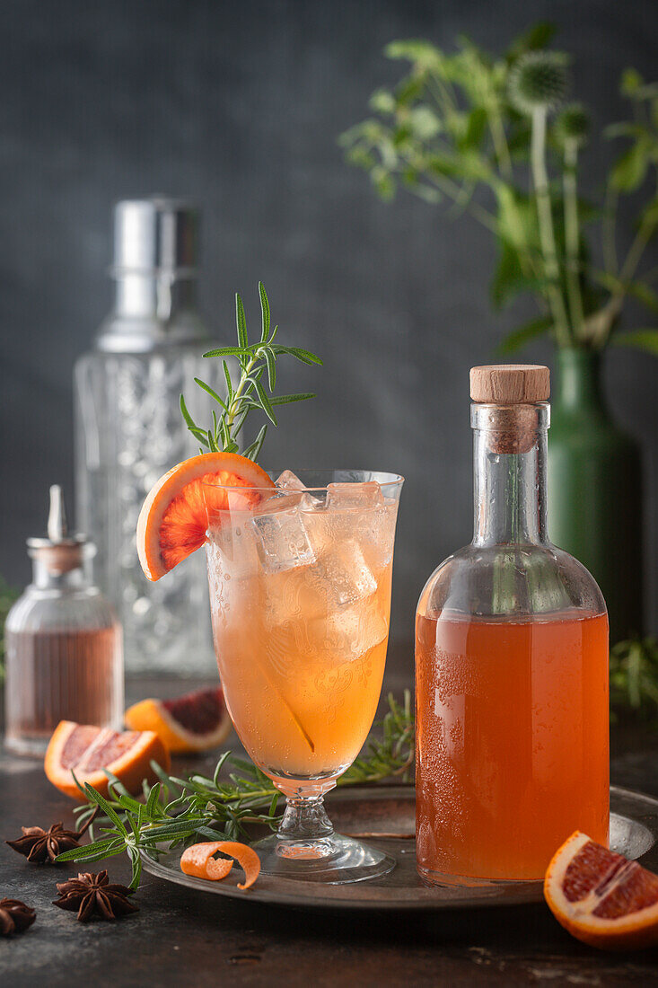 Colourful cocktail on ice with blood orange syrup and club soda on ice in a vintage glass with syrup bottle, citrus fruits and rosemary garnish