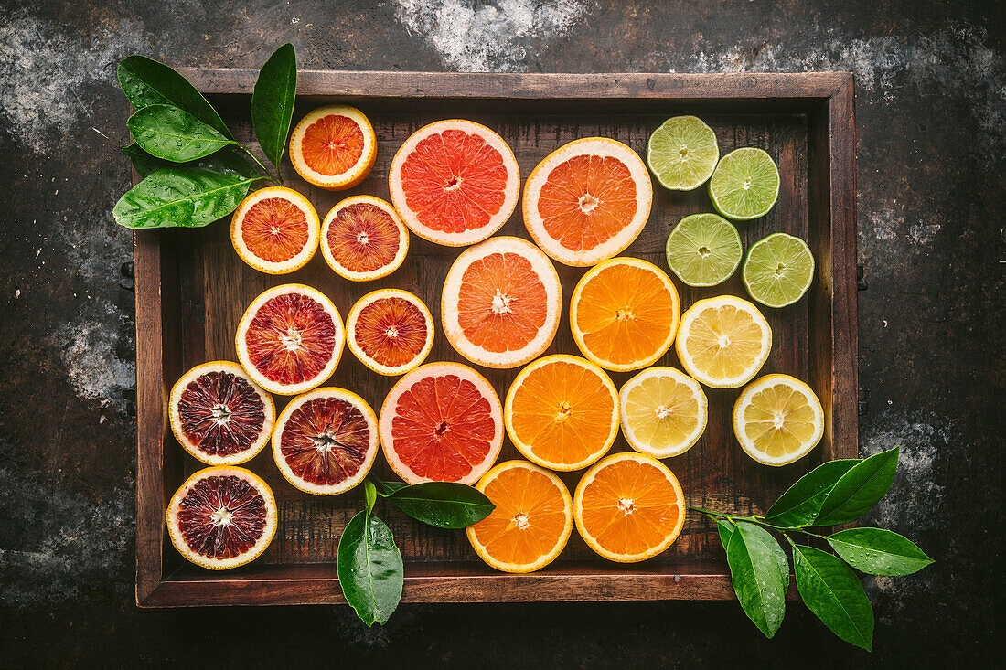 Citrus fruits (orange, blood orange, grapefruit, lemon, lime) halved and sorted by colour in a rustic wooden box with fresh citrus leaves