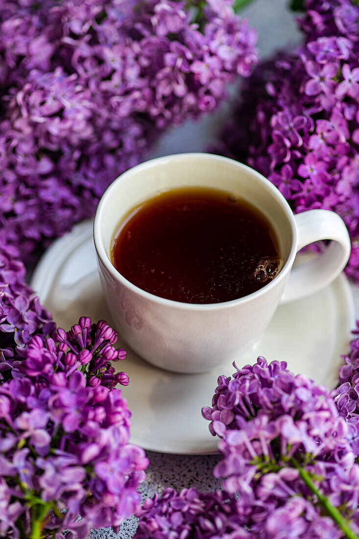 From above white cup of tea and beautiful lilac flowers around on grey concrete background