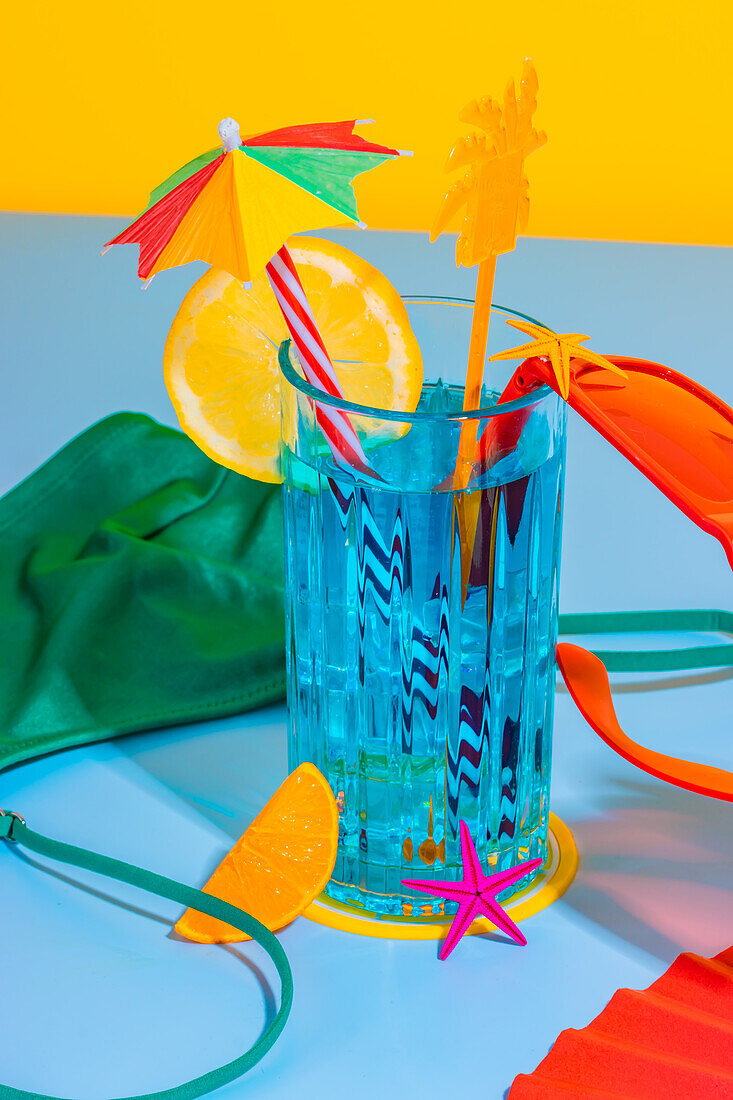 Komposition aus Cocktailglas und aufgeschnittener Orangenfrucht mit Strohhalm, roten Gläsern auf der Oberfläche mit innerer Abnutzung, Handfächer auf blauem und gelbem Hintergrund