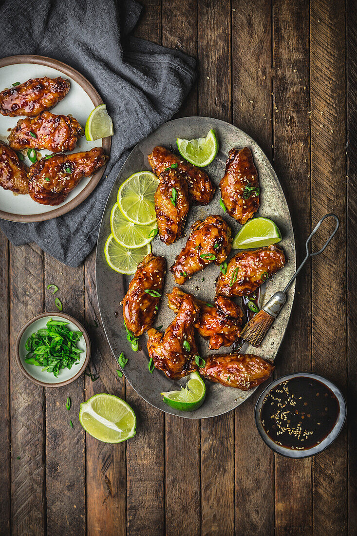 Glazed chicken wings on metal plate and serving platter with limes, sauce and spring onions