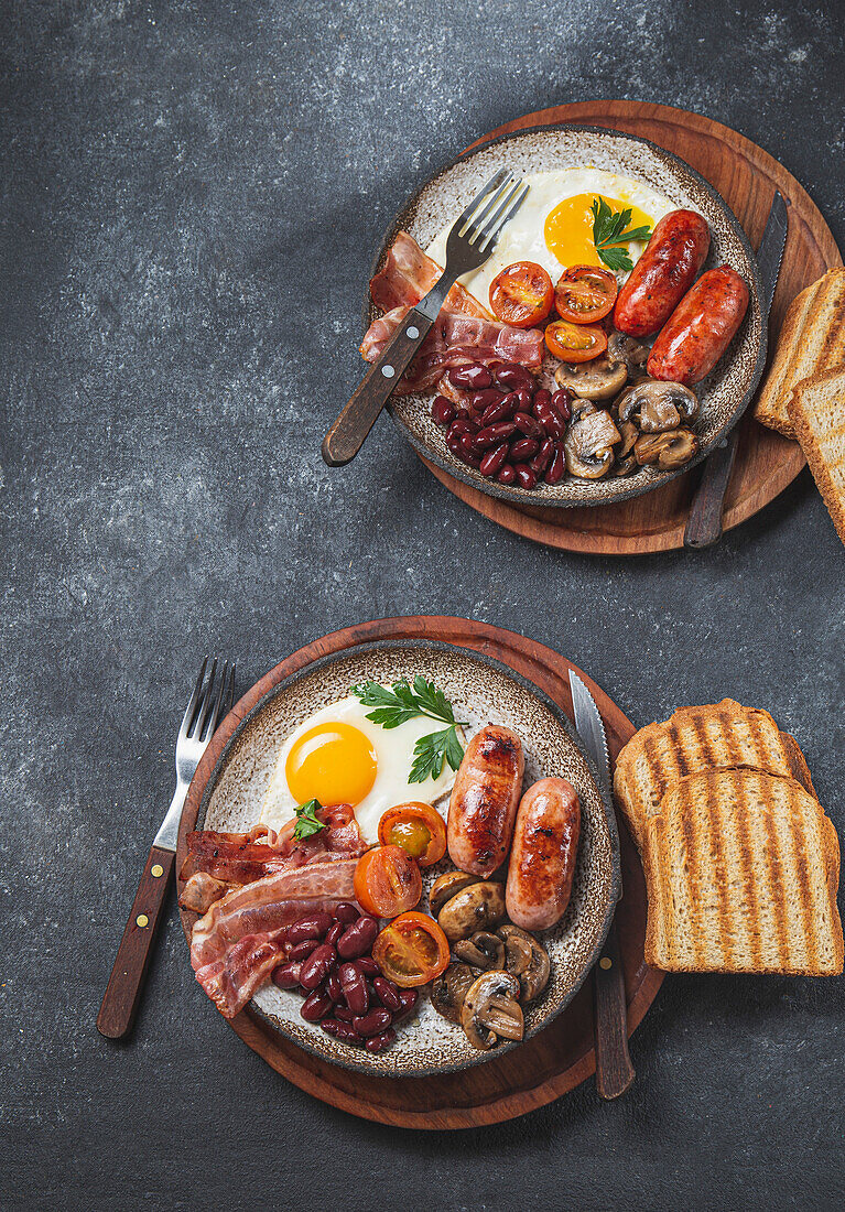 Traditional full English breakfast with fried eggs, sausages, beans, mushrooms, grilled tomatoes, bacon and toasts on gray plates