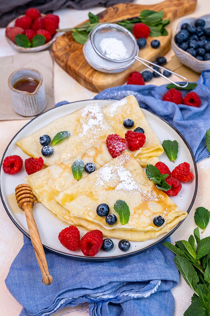Crepes garnished with blueberries, raspberries, fresh mint leaves and icing sugar on a round plate