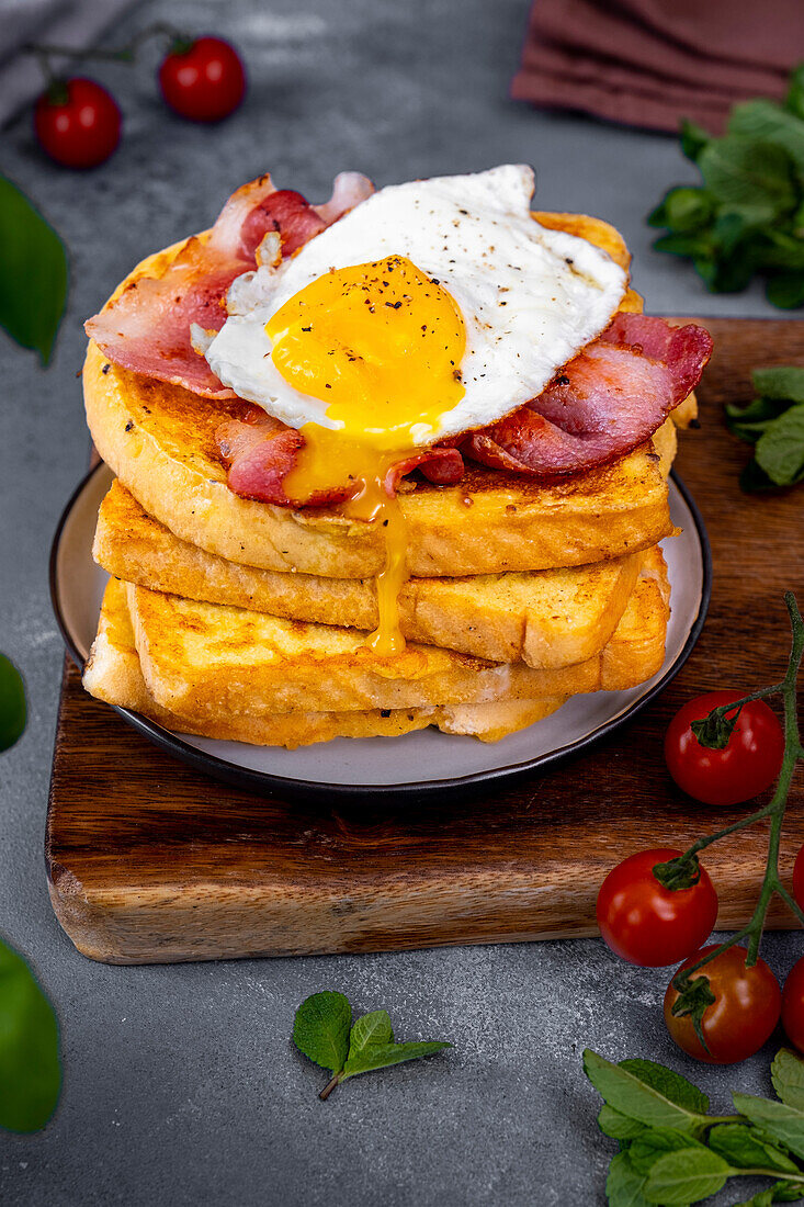 A stack of egg-covered slices of bread topped with fried bacon and egg. The egg yolk is dripping