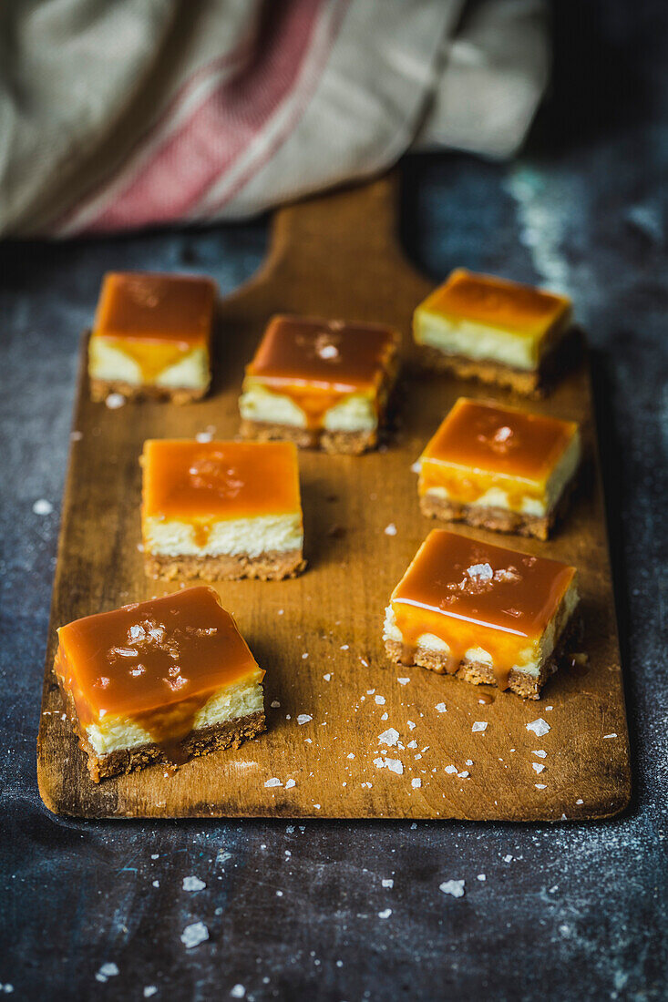 Small squares of cheescake topped with caramel sauce and sea salt on a wooden board