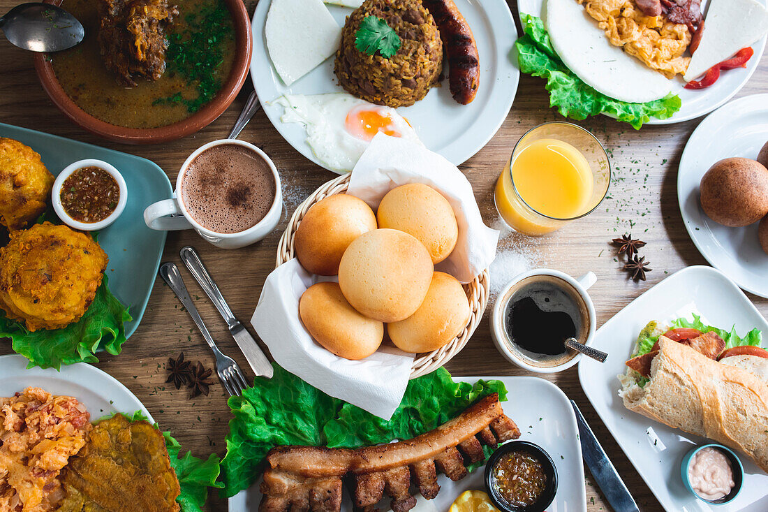 Top view of an array of appetising Colombian dishes with papas rellenas pan de bono chicharron bunuelos and caldo de costilla with various drinks on a wooden table in a restaurant