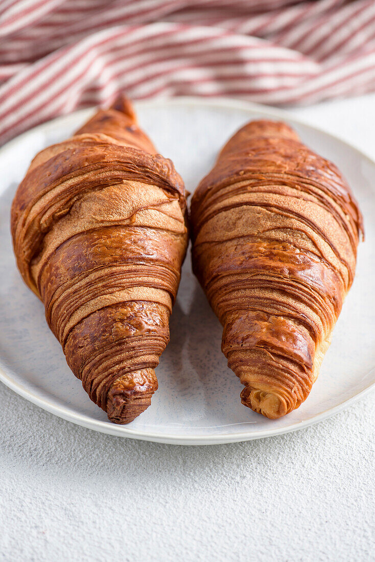 Two croissants on a white plate in front of a red striped kitchen towel