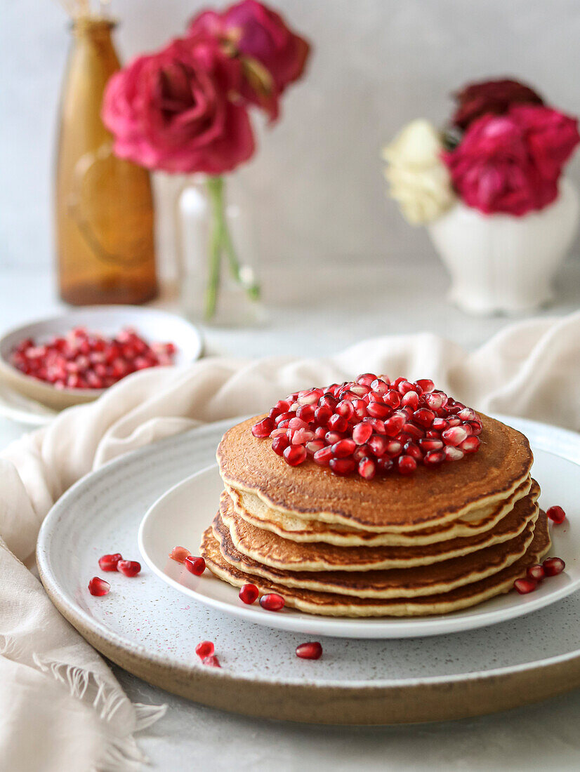 Stapel Pfannkuchen mit Granatapfel und roten Blumen auf hellem Hintergrund