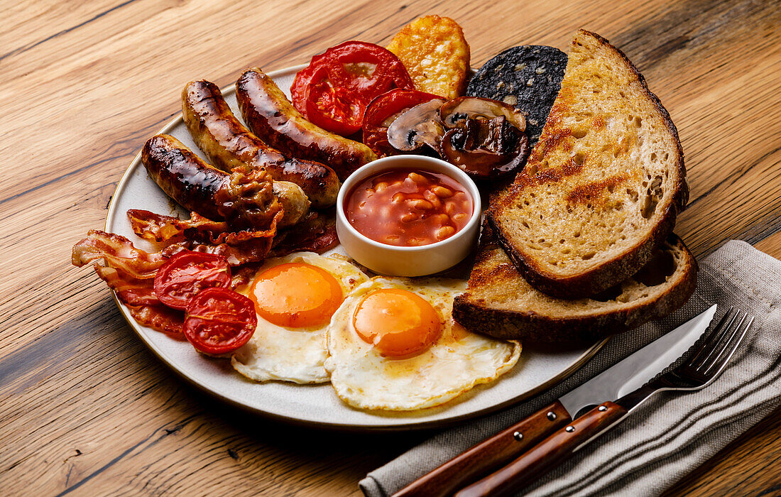 A full English breakfast with fried eggs, sausages, bacon, black pudding, beans and toast on a wooden backdrop