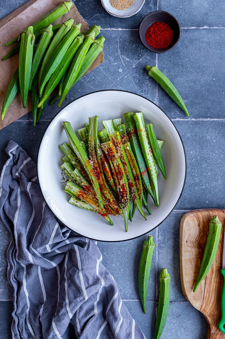 Okra pods sliced lengthwise in a white bowl, spices on them, whole okra pods around.