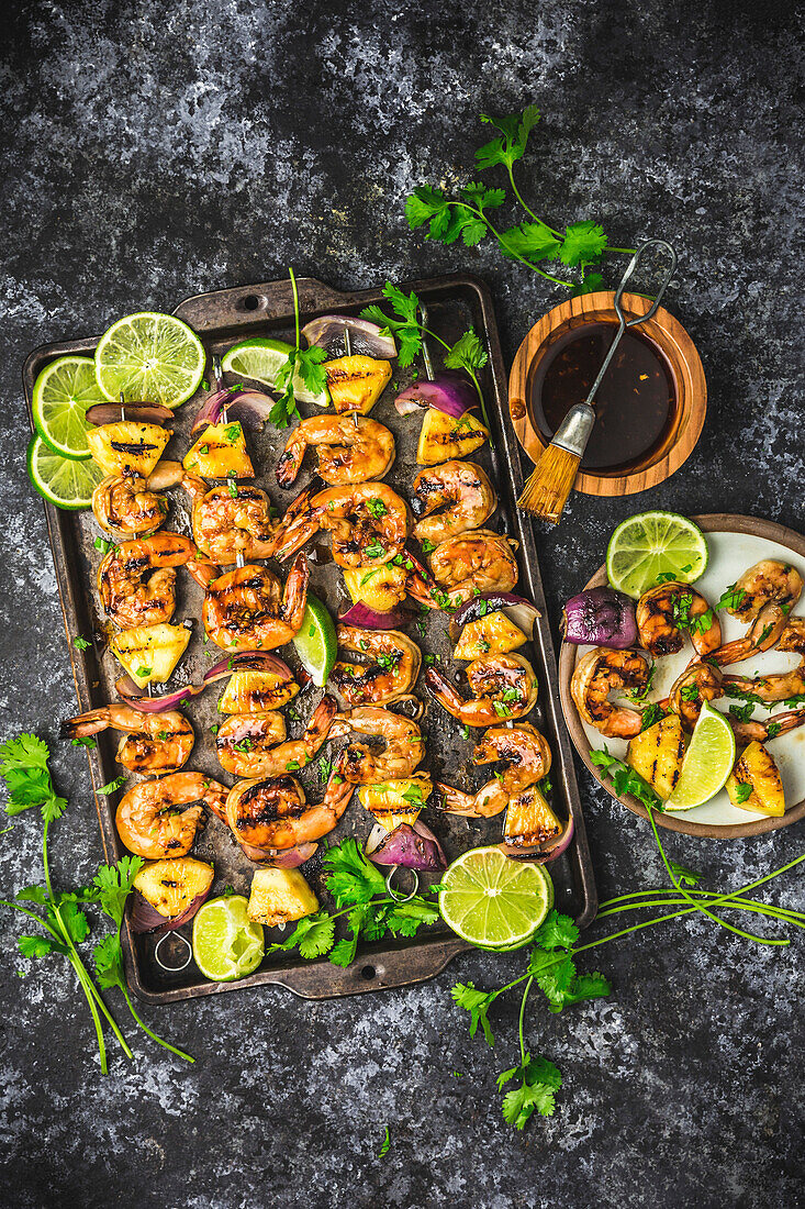 Skewers with grilled prawns, pineapple and red onions on a baking tray and plate with glaze in a bowl