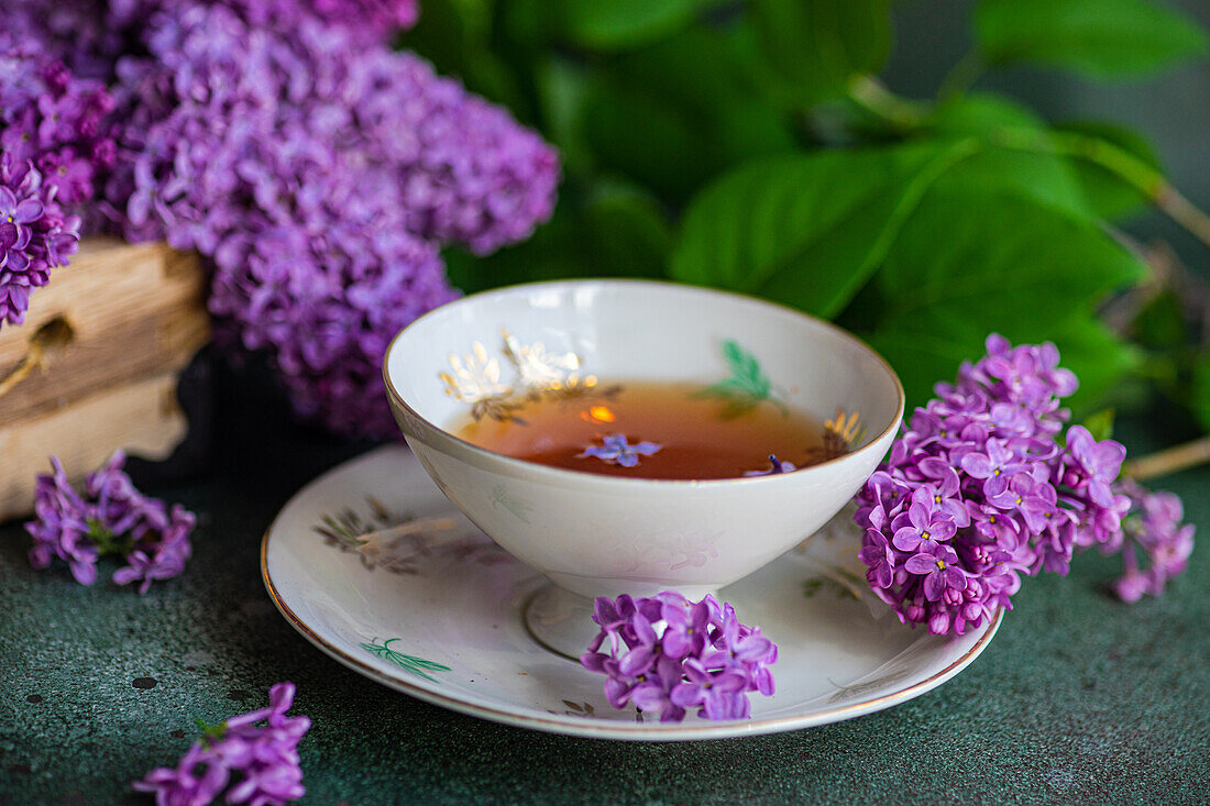 Front view of a delicious black tea in a white vintage cup on a mint green concrete table with aromatic lilac flowers