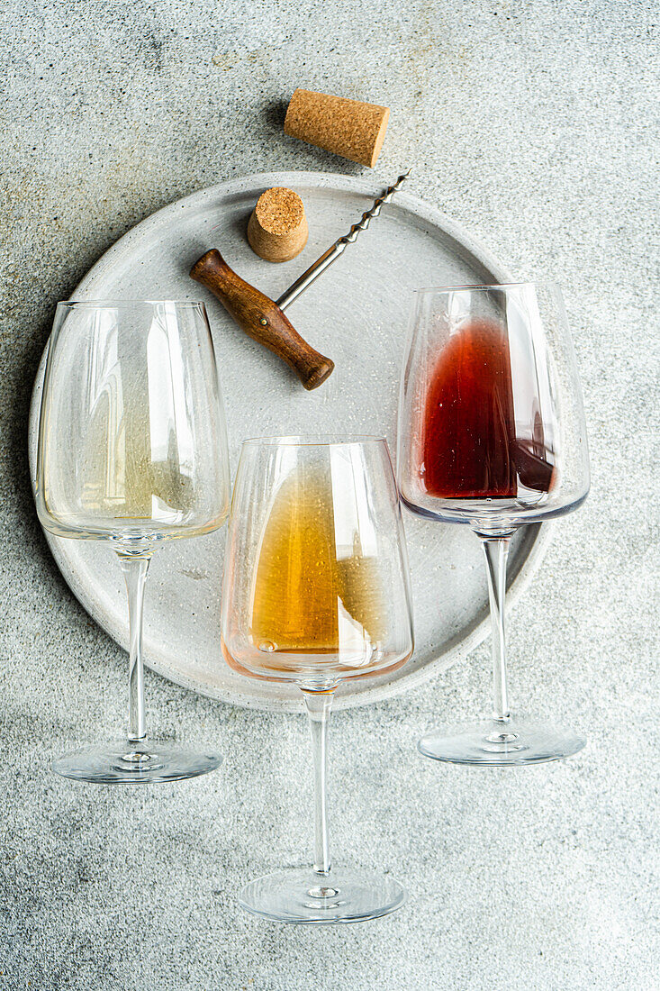 High angle of three varieties of Georgian dry wine glasses (white, amber and red) lying on a plate with a corkscrew and a cork on concrete grey table