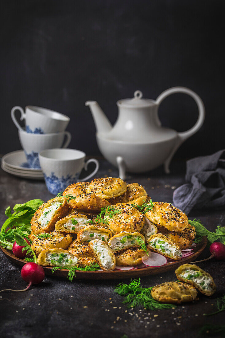 Eine Platte mit mit Käse gefüllten Gougeres, die mit allem Möglichen gewürzt sind, mit einer Teekanne im Hintergrund