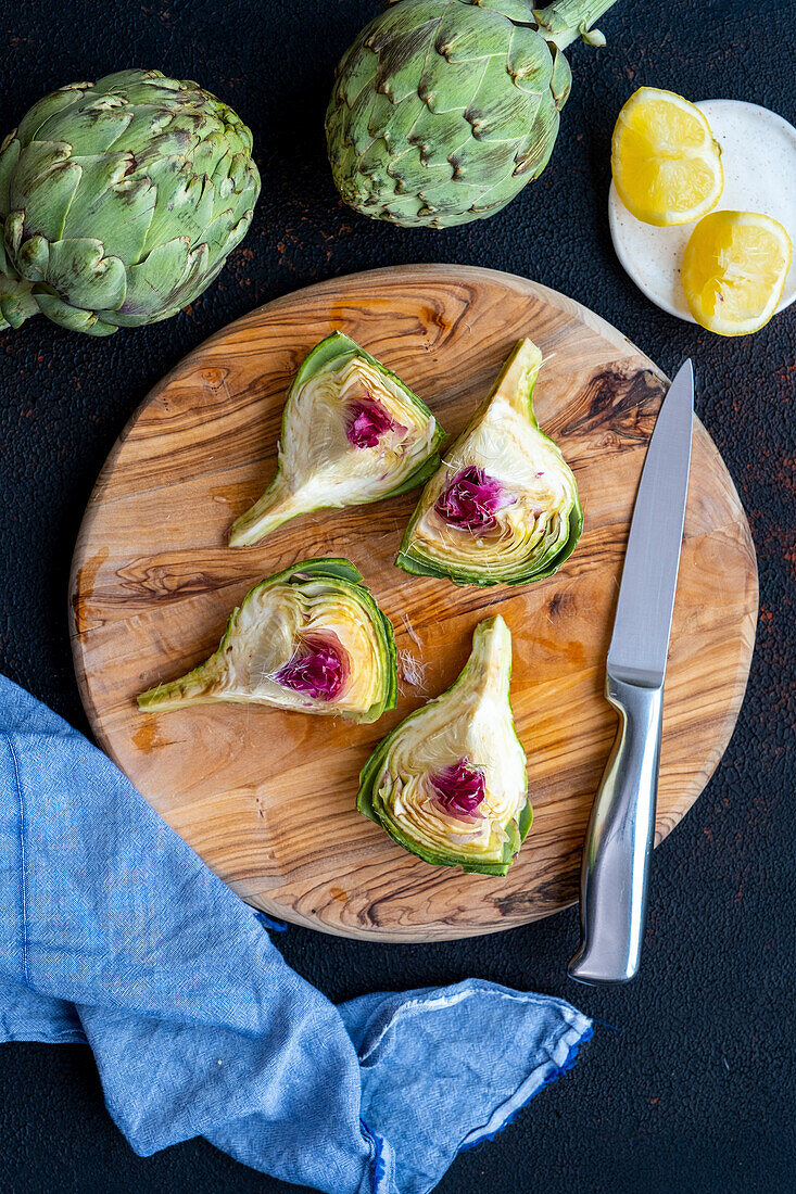 An artichoke quartered on a wooden board, accompanied by a knife, lemon slices and whole artichokes
