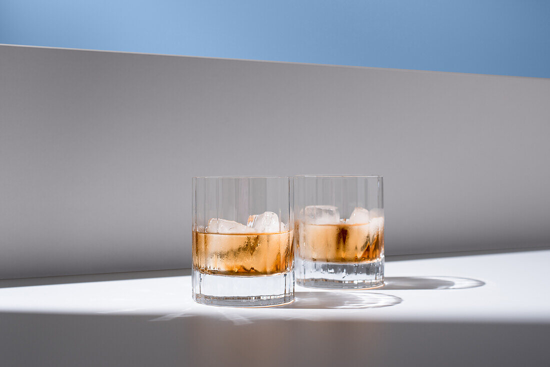 Transparent glasses filled with cold, refreshing Scotch whiskey with ice cubes on a white surface in front of a white wall