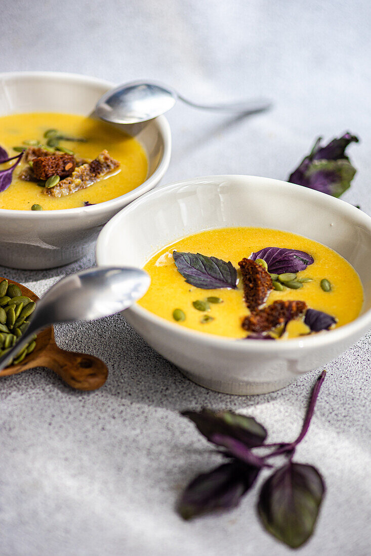 Front view of cropped bowls with pumpkin cream soup with basil herb, rye bread and seeds on blurred grey background with leaves