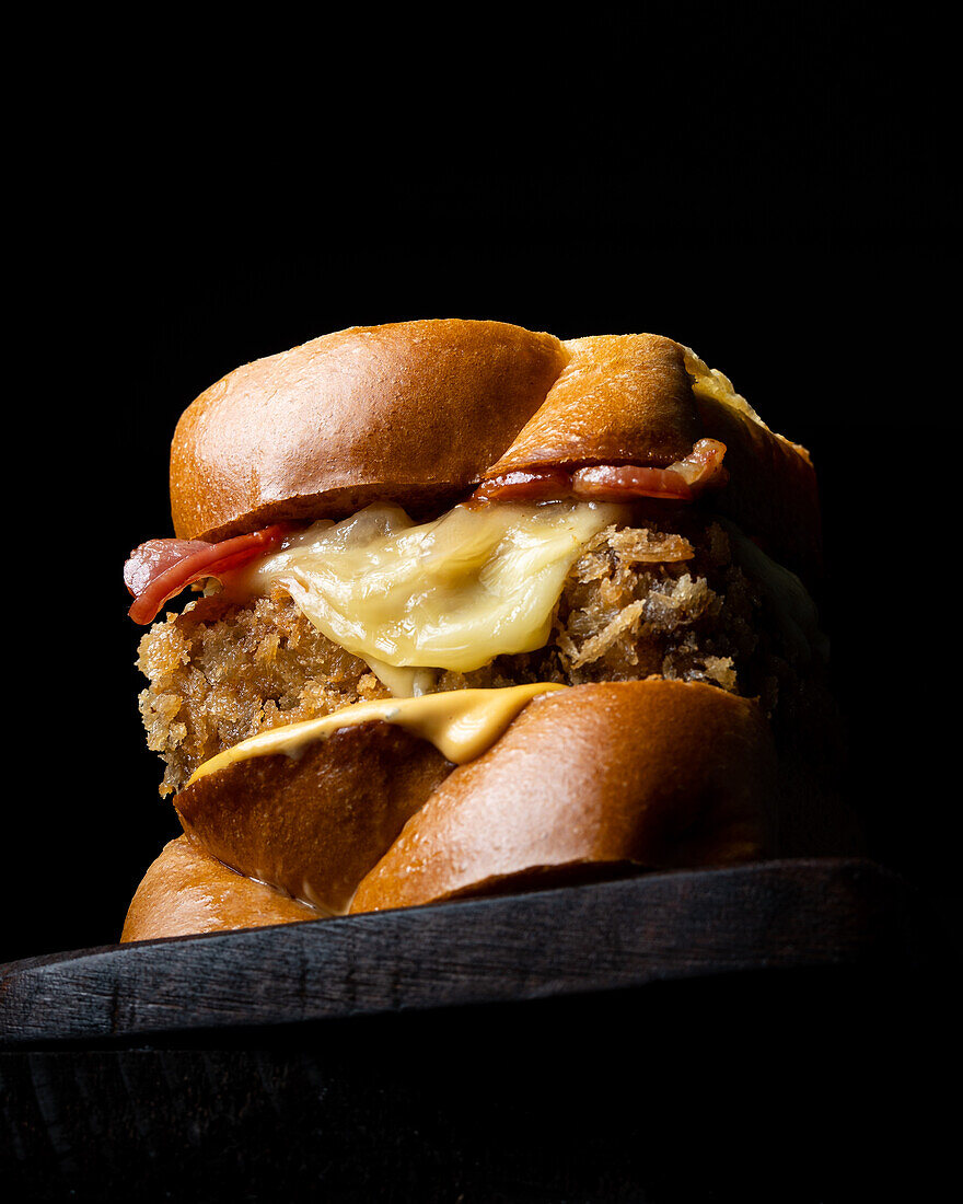 From below appetising burger with fresh buns, cheese and bacon served on a wooden board on a black background