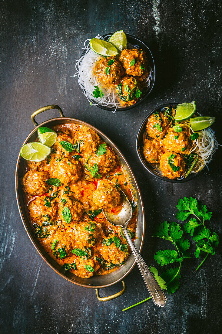 Fleischbällchen in Currysauce in Servierschalen mit Nudeln und Limettenscheiben